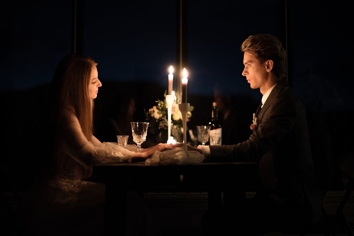 Side view of loving young couple in elegant outfits holding hands while sitting in front of each other during romantic dinner with burning candles in restaurant